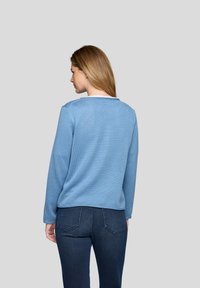 Frau mit glattem blonden Haar, die ein strukturiertes, hellblaues Langarmoberteil und dunkelblaue Jeans trägt und mit dem Rücken zu einem schlichten grauen Hintergrund steht.