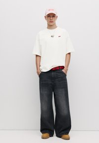 Overdimensioneret hvid t-shirt med rød logo, løse grå jeans, rød mønstret bælte, beige sko og en lyserød kasket med broderede detaljer.