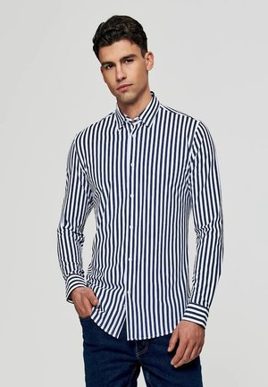 Mężczyzna ubrany w granatowo-białą koszulę w pionowe paski na guziki z długim rękawem oraz ciemnoniebieskie dżinsy, pozujący z jedną ręką w kieszeni.