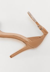 Béžová sandálová obuv na vysokém podpatku s elegantním designem, vybavená páskem zdobeným kamínky a štíhlým podpatkem. Hladký syntetický materiál.