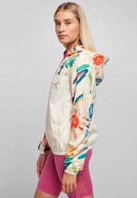 Lightweight, cream jacket with colorful floral patterns on sleeves, elastic cuffs, and a hood. Pocket on front and zipper closure.