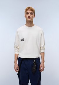 Κρεμ μπλουζάκι με μακριά μανίκια, μαύρο λογότυπο στη αριστερή πλευρά και πορτοκαλί ραφές στους ώμους, συνδυασμένο με ναυτικό μπλε παντελόνι και μαύρο αξεσουάρ.