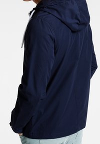 Granatowa lekka kurtka z kapturem, białym sznurkiem i gładką fakturą. Charakteryzuje się luźnym krojem i minimalistycznymi przeszyciami.