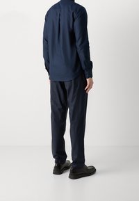 Marinblå långärmad skjorta med ett bakveck, i kombination med löst sittande marinblå byxor och svarta skor med en texturerad yta.