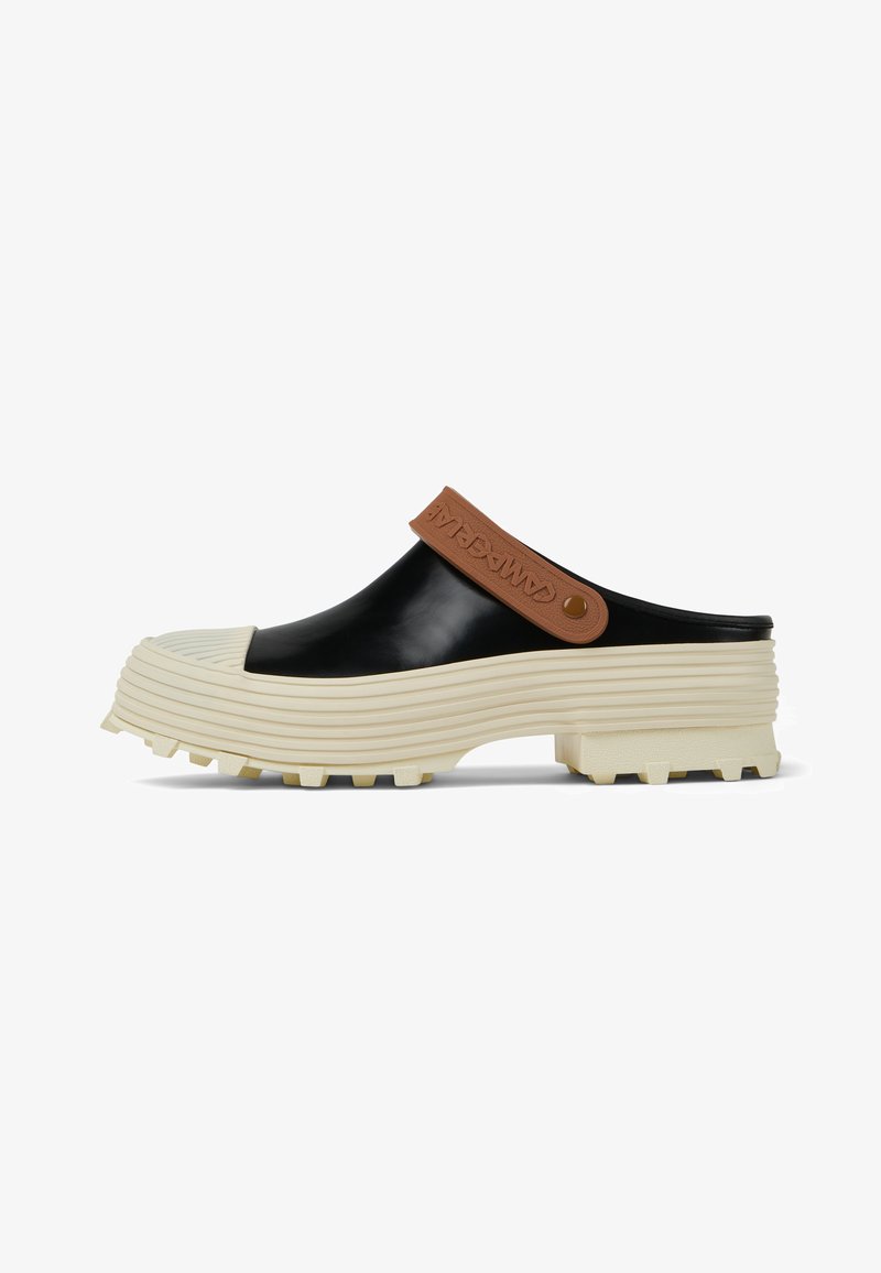 Black rubber clogs with a beige ridged outsole, featuring a brown strap with embossed logo detail. Chunky and textured design, open-backed.