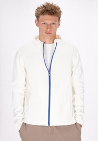 Witte zip-up jas met een blauwe rits, voorzien van een hoge kraag en twee zijzakken, gedragen over een lichtgekleurde blouse. Gladde textuur.