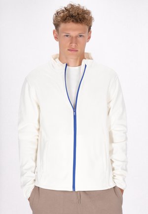 Hvid zip-up jakke med en blå lynlås, der har en høj krave og to sidelommer, båret over en lysfarvet skjorte. Glat tekstur.