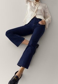 Mörkblå utsvängda jeans i denim, kombinerade med en krämfärgad silkesblus med knytkrage, och svarta loafers med guldförzieringar.