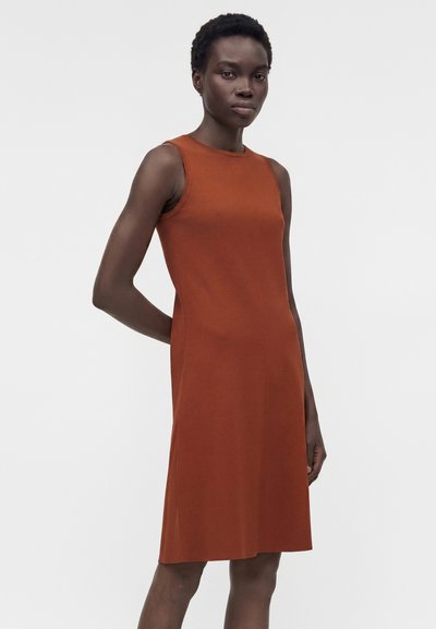 Γυναίκα με κοντά, σγουρά μαλλιά που φοράει ένα αμάνικο, φορέμα μήκους μέχρι το γόνατο σε σκουρόχρωμο ρουστίκ χρώμα, στέκεται μπροστά σε ένα απλό ανοιχτόφωτο φόντο.