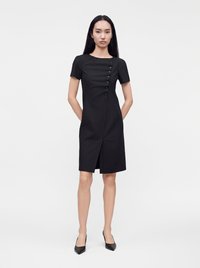 Femme debout sur un fond uni, portant une robe noire moulante longueur genou avec des boutons latéraux et une fente sur le devant, assortie à des talons noirs.