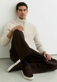 Krem pulover z teksturirano pletenino, visokim ovratnikom in zadrgo. Združeno z širokimi rjavimi hlačami in temnimi supergami, sedi ob svetlem zidu.