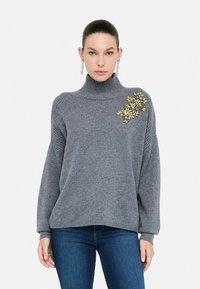 Vrouw in een grijze coltrui met gouden bloemenversiering op de schouder en blauwe jeans, recht tegen een witte achtergrond gekeerd.