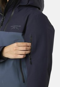 Vandtæt jakke med et navy- og blåfarvet blokdesign, med lynlåslommer og justerbar hætte. Glat tekstur, funktionelle detaljer.