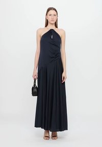 Temno modra maksi obleka s halter ovratnikom in zvitim sprednjim delom, tekoč material in asimetrično drapiranje, v kombinaciji z majhno črno torbico.