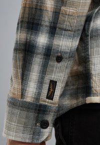 Camisa de cuadros de manga larga en una mezcla de colores azul marino, beige y gris, con botones negros y una etiqueta de logo cerca del puño.