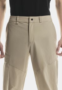 Hombre vestido con pantalones cargo beige y una camiseta negra, mostrado desde la cintura hasta la mitad del muslo, de pie con las manos relajadas a los lados.
