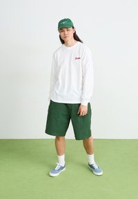 Long-sleeve white shirt with red logo, green shorts, green cap, blue patterned sneakers, and white crew socks on green floor.