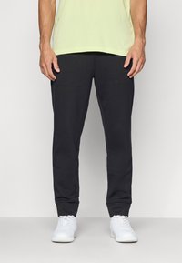 Dunkelgraue Jogginghosen mit weicher Textur, schmaler Passform und elastischen Bündchen, kombiniert mit weißen Sneakers und einem hellgelben Shirt.
