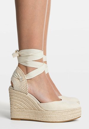 Beige espadrille kilehælssandaler med ankelbind, båret på bare fødder mod en hvid baggrund.