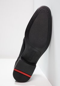 Chaussure noire avec une tige texturée et une semelle en caoutchouc lisse, ornée d'une bande rouge sur le talon et d'un motif en zigzag sur le bord.