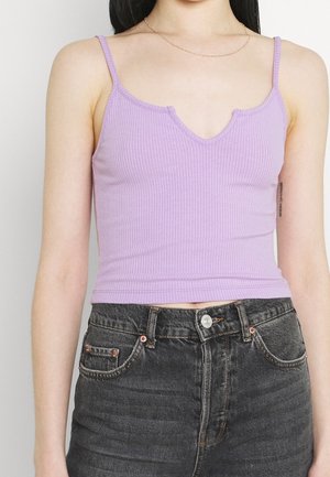Kobieta ubrana w lawendowy, prążkowany, bezrękawnik z głębokim dekoltem w serek oraz czarne, wysokowystające dżinsy z miedziowanymi nitami.