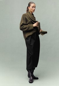 Übergroßer, gerippter olivgrüner Pullover mit einem drapierten Ausschnitt, kombiniert mit hoch taillierten schwarzen Hosen und knöchellangen schwarzen Stiefeletten.
