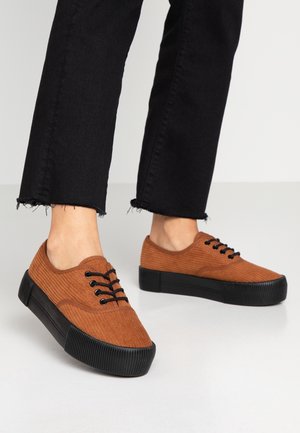 Persona que lleva zapatillas de pana marrón con suelas negras gruesas y cordones negros, combinadas con pantalones negros recortados con dobladillos deshilachados.