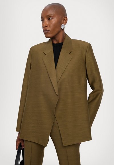 Blazer oversized de culoare verde măslin, cu un design structurat, material texturat, rever ascuțit și închidere cu un singur nasture, asortat cu un top negru.