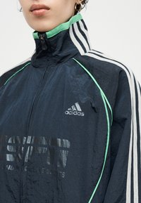 Κοντινό πλάνο ενός ατόμου που φοράει μια μπλε σκούρα αθλητική ζακέτα Adidas με πράσινη περίμετρο, λευκές ρίγες στα μανίκια και κλειστό φερμουάρ στο ψηλό γιακά.