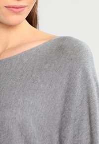 Lysegrå strikket sweater med en vid halsudskæring, der fremhæver en blød tekstur og subtil ribbet mønster, der hænger blidt over skuldrene.