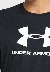 Camiseta de algodón negra con un gran logo blanco de Under Armour y texto. El cuello redondo es acanalado y la tela tiene una textura suave.