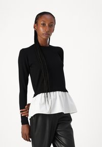 Maglione nero a maglia con maniche lunghe, caratterizzato da un orlo peplum arricciato bianco. Il modello indossa pantaloni in pelle neri. Texture liscia, design aderente.
