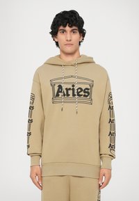 Bež dukserica napravljena od mekanog materijala, središnji crni grafički ispis na kojem piše "Aries" i ponovljeni logotipi duž rukava. Uključene su šnurice.