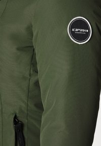 Chaqueta aislante verde con una textura suave. Presenta un parche de logo redondo en blanco y negro en la parte superior del brazo y un bolsillo lateral con cremallera.