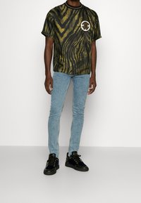 Camiseta con rayas de tigre en verde y negro, con cuello redondo y parche con logo en el pecho, combinada con vaqueros pitillo azul claro y zapatillas negras.