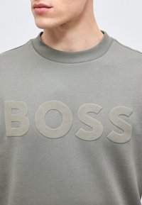 Κοντινό πλάνο ενός ατόμου που φοράει ανοιχτό γκρι φούτερ με μεγάλους υφασμάτινους χαρακτήρες που σχηματίζουν τη λέξη "BOSS" στο στήθος.