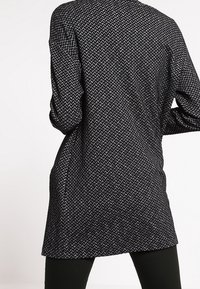 Dlouhý kardigan s černo-bílým vzorem houndstooth, rovného střihu a texturovaného materiálu, s dlouhými rukávy a uvolněným střihem.