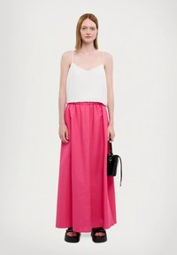 Top bianco con spalline sottili, abbinato a una lunga gonna rosa con vita elasticizzata. La modella tiene una piccola borsa nera e indossa sandali con plateau.