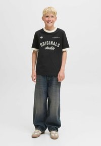Camiseta negra con texto blanco "ORIGINALS studio" y cuello crema; jeans de pierna ancha de mezclilla; zapatos en tonos claro y crema. Fondo liso.