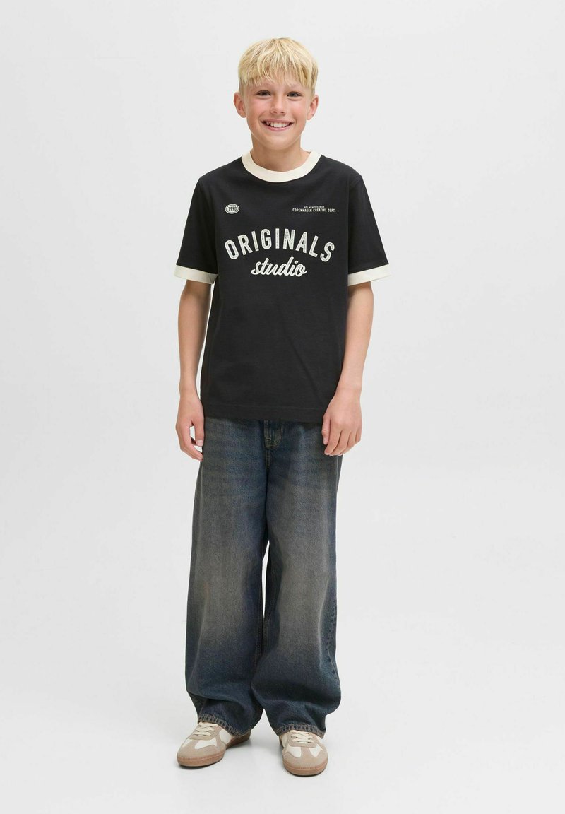 Camiseta negra con texto blanco "ORIGINALS studio" y cuello crema; jeans de pierna ancha de mezclilla; zapatos en tonos claro y crema. Fondo liso.
