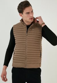 Vesta umflată maro cu un design pătrățos vertical, închidere cu fermoar, guler înalt și buzunare laterale. Purtați peste un tricou negru cu mâneci lungi.
