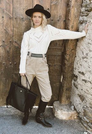 Weißer Strickpullover mit Ausschnitten, beige hochtaillierte Hose, schwarze Handtasche mit Fransen und knielangen schwarzen Stiefeln. Schwarzer Hut mit breiter Krempe.