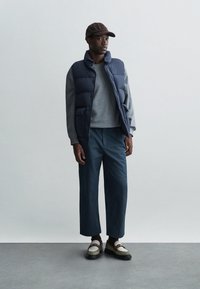Chaleco acolchado navy sobre un suéter gris, combinado con pantalones anchos azules y zapatos texturizados en crema y marrón. Una gorra marrón completa el atuendo.