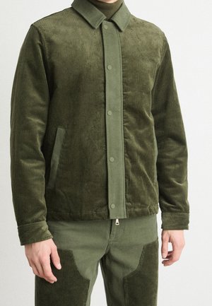 Homme portant une veste en velours côtelé vert olive et un pantalon assorti avec des boutons-pression et des poches latérales, debout devant un fond blanc uni.