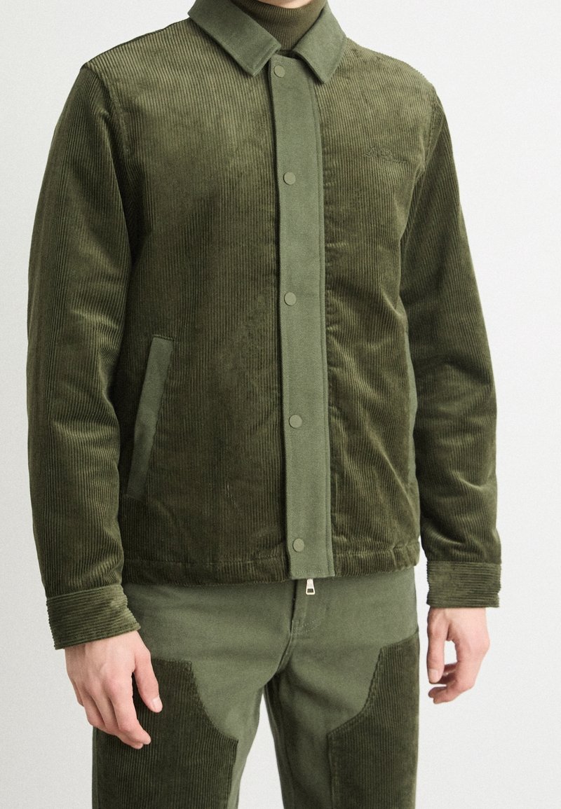 Homme portant une veste en velours côtelé vert olive et un pantalon assorti avec des boutons-pression et des poches latérales, debout devant un fond blanc uni.