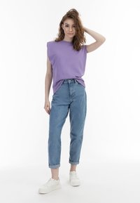 Lyse lilla ærmeløs top, blå højtaljede jeans og hvide sneakers. Model poserer med hånden i håret foran en ensfarvet hvid baggrund.