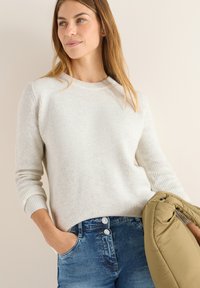 Hellgrauer gerippter Pullover mit Rundhalsausschnitt; das Modell hält eine olivgrüne Daunenjacke. Blaue Jeans mit sichtbarem Doppelknopfverschluss.