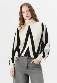 Svart-hvitt mønstrete genser med høy krage, med sikksakk-design. Kombinert med lysegrå jeans for et kontrastfylt utseende.