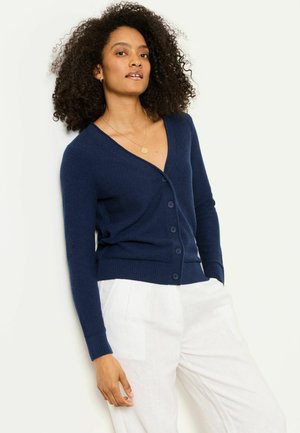 Woman with curly hair wearing a navy blue buttoned cardigan and white pants, posing against a light background.