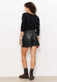 Un pull en tricot noir, un short en cuir noir avec taille élastique, et des bottines en daim marron avec semelles noires, présentés sur un fond neutre.